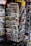 A rack filled with a variety of newspapers and magazines, featuring multiple languages and diverse topics. The publications are neatly arranged in several tiers, showcasing different headlines and imagery. The setting appears to be a newsstand, possibly located in a public area with other items partially visible in the background.