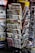 A rack filled with a variety of newspapers and magazines, featuring multiple languages and diverse topics. The publications are neatly arranged in several tiers, showcasing different headlines and imagery. The setting appears to be a newsstand, possibly located in a public area with other items partially visible in the background.