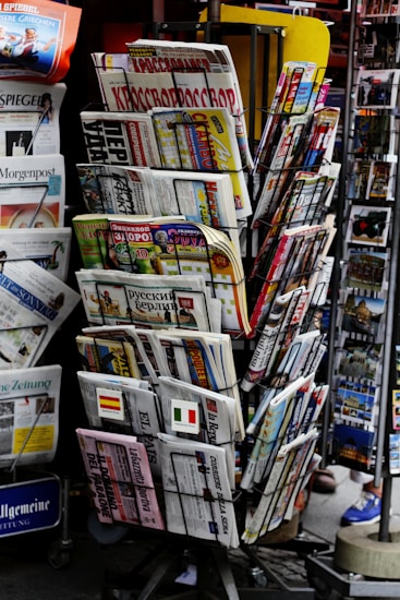 A rack filled with a variety of newspapers and magazines, featuring multiple languages and diverse topics. The publications are neatly arranged in several tiers, showcasing different headlines and imagery. The setting appears to be a newsstand, possibly located in a public area with other items partially visible in the background.