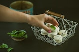 A hand is reaching for a macaron with a mint leaf on top from a wire basket filled with similar treats. A small bowl of fresh mint leaves is placed nearby, and a vintage tin can is in the background.
