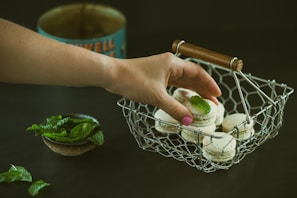 A hand is reaching for a macaron with a mint leaf on top from a wire basket filled with similar treats. A small bowl of fresh mint leaves is placed nearby, and a vintage tin can is in the background.