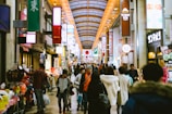 A busy indoor shopping arcade with numerous people walking and shopping. Brightly lit signs with Japanese characters hang above, and various storefronts display colorful products. A large Japanese flag is displayed prominently in the distance, contributing to a bustling and lively atmosphere.