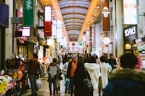 A busy indoor shopping arcade with numerous people walking and shopping. Brightly lit signs with Japanese characters hang above, and various storefronts display colorful products. A large Japanese flag is displayed prominently in the distance, contributing to a bustling and lively atmosphere.