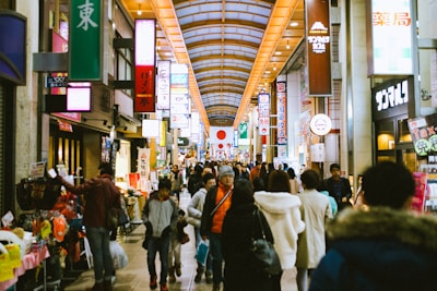 A busy indoor shopping arcade with numerous people walking and shopping. Brightly lit signs with Japanese characters hang above, and various storefronts display colorful products. A large Japanese flag is displayed prominently in the distance, contributing to a bustling and lively atmosphere.