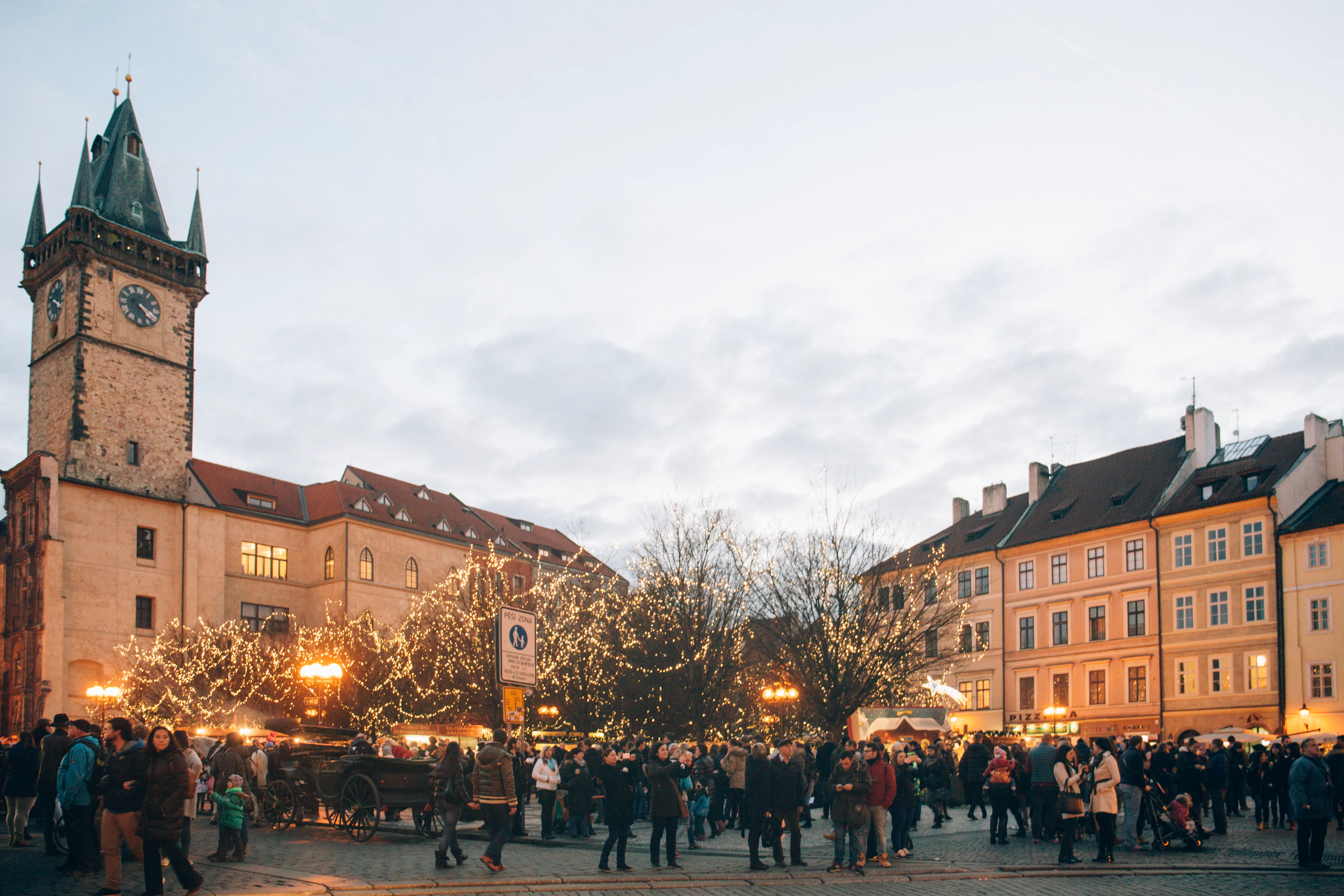 Old Town Square's Christmas market glows with string lights and crowds at dusk beneath the clock tower.