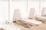 A row of white plastic lounge chairs is placed on a sunlit patio, casting shadows on the ground. The setting is minimalist and bright, with a white lattice fence in the background adding to the clean and serene atmosphere.