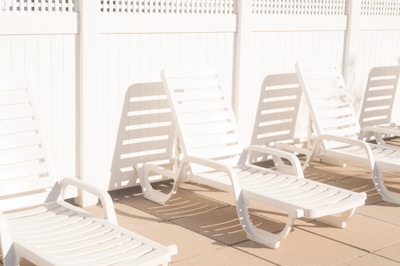 A row of white plastic lounge chairs is placed on a sunlit patio, casting shadows on the ground. The setting is minimalist and bright, with a white lattice fence in the background adding to the clean and serene atmosphere.