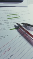 Several sheets of paper are spread out with handwritten notes and printed text. Two pens are placed on top, one with a black tip and the other with a red tip. Some sections of the printed text are highlighted in green.