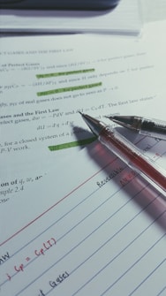 Several sheets of paper are spread out with handwritten notes and printed text. Two pens are placed on top, one with a black tip and the other with a red tip. Some sections of the printed text are highlighted in green.