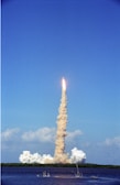 A rocket is launching into a clear blue sky with a thick trail of smoke stretching from the ground to the rocket. The launch site is located near a body of water, with some foliage visible in the distance. Puffs of smoke linger around the launch pad.