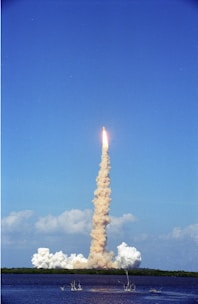 A rocket is launching into a clear blue sky with a thick trail of smoke stretching from the ground to the rocket. The launch site is located near a body of water, with some foliage visible in the distance. Puffs of smoke linger around the launch pad.