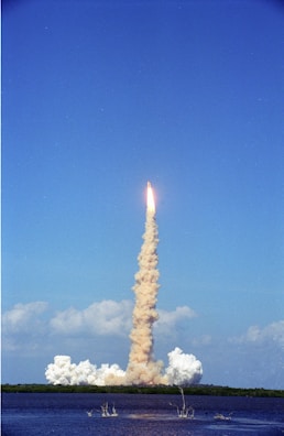 A rocket is launching into a clear blue sky with a thick trail of smoke stretching from the ground to the rocket. The launch site is located near a body of water, with some foliage visible in the distance. Puffs of smoke linger around the launch pad.