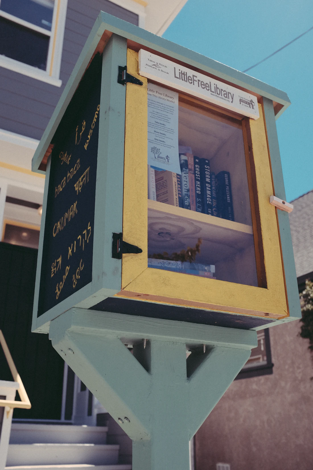 A classic blue and yellow little free library on a post with a welcome sign