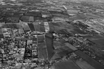 An aerial view of a landscape depicting a patchwork of rectangular fields and farmland. Roads and small clusters of buildings are interspersed throughout the area. The horizon shows a dense collection of structures indicating a more urban area. The image is in black and white, with various shades of gray highlighting the contrast between different plots of land and structures.