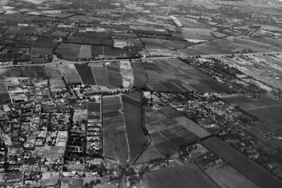 An aerial view of a landscape depicting a patchwork of rectangular fields and farmland. Roads and small clusters of buildings are interspersed throughout the area. The horizon shows a dense collection of structures indicating a more urban area. The image is in black and white, with various shades of gray highlighting the contrast between different plots of land and structures.