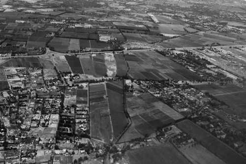 An aerial view of a landscape depicting a patchwork of rectangular fields and farmland. Roads and small clusters of buildings are interspersed throughout the area. The horizon shows a dense collection of structures indicating a more urban area. The image is in black and white, with various shades of gray highlighting the contrast between different plots of land and structures.