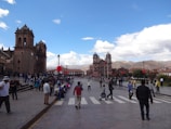 An expansive plaza is bustling with people, with a prominent and ornate church on the left side. The sky is bright with scattered clouds, and red decorations, possibly for a celebration, are visible. The backdrop features mountains in the distance. The scene conveys a sense of activity and community engagement.
