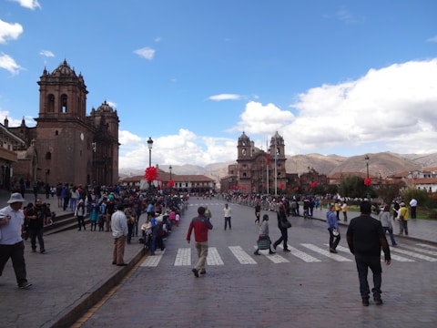 An expansive plaza is bustling with people, with a prominent and ornate church on the left side. The sky is bright with scattered clouds, and red decorations, possibly for a celebration, are visible. The backdrop features mountains in the distance. The scene conveys a sense of activity and community engagement.