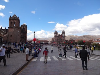 An expansive plaza is bustling with people, with a prominent and ornate church on the left side. The sky is bright with scattered clouds, and red decorations, possibly for a celebration, are visible. The backdrop features mountains in the distance. The scene conveys a sense of activity and community engagement.