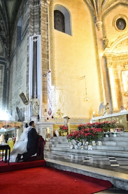 A couple kneels on a red carpet in a grand church interior, beautifully adorned with statues, religious symbols, and floral arrangements. The intricate architecture features high ceilings, arched windows, and ornate decorations, creating a solemn and sacred atmosphere. A priest stands at the altar conducting a religious ceremony.