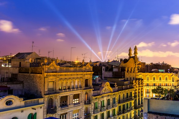 A night scene featuring a cityscape with brightly lit, historic buildings adorned with architectural details. The sky is a vibrant mix of deep blue and pink hues, illuminated by multiple searchlights beaming across the skyline.