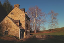 A rustic stone house with a pitched roof sits in a serene rural setting. The house is surrounded by leafless trees with a clear blue sky in the background. There is a grassy field leading up to the house, creating a peaceful and isolated atmosphere.
