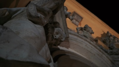 A close-up view of a stone or marble sculpture featuring expressive human faces set against an architectural backdrop with columns and intricate detailing. The scene is dimly lit, suggesting evening or nighttime, with a warm light illuminating the details.