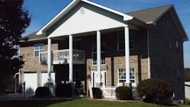 A two-story house with a brick facade, white columns, and a porch with a white railing. The house has multiple windows and a prominent gable roof. In the foreground, there is a well-manicured lawn and some bushes. A tree partially obscures the left side of the house.