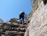 A mountaineer wearing an Amadou LLC backpack climbing a rocky trail under a clear blue sky