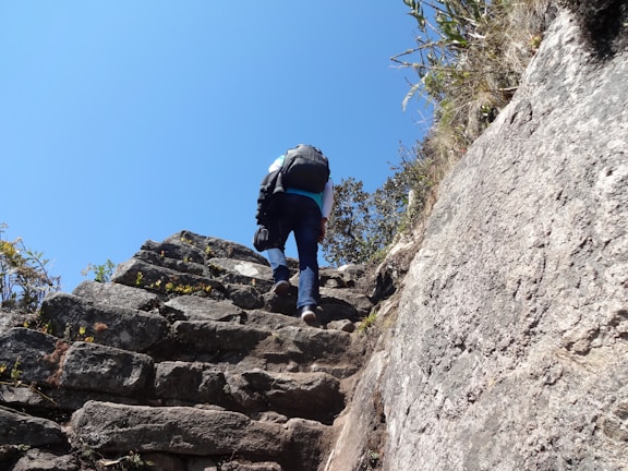 A mountaineer wearing an Amadou LLC backpack climbing a rocky trail under a clear blue sky