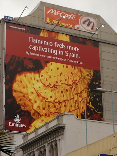 A large outdoor advertisement on the side of a building promoting travel to Spain by Emirates. The focus of the advertisement is on flamenco, symbolized by a vivid image of a traditional flamenco dress in yellow and red colors. Above the advertisement, there is another ad for a McCafé, indicating its location at 267 Queen St.