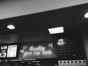 A black and white photograph of a fast-food restaurant interior with a neon sign reading 'Quality you can taste.' The walls are tiled, and a menu board is visible displaying prices for various food items.