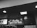 A black and white photograph of a fast-food restaurant interior with a neon sign reading 'Quality you can taste.' The walls are tiled, and a menu board is visible displaying prices for various food items.
