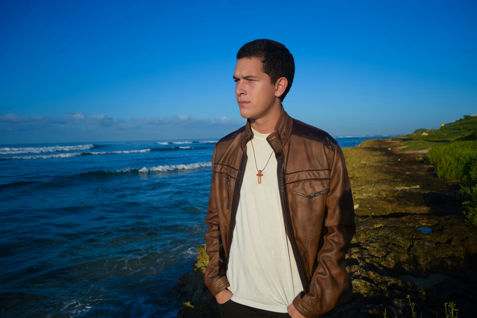 A person stands on a rocky shoreline with waves in the background. They are wearing a brown leather jacket, white shirt, and a necklace with a cross. The setting sun casts a warm glow over the scene.