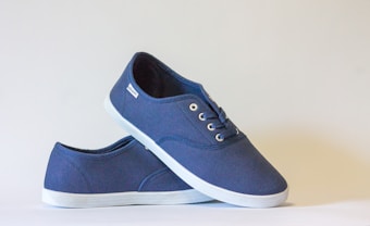 A pair of dark blue canvas sneakers with white rubber soles. One shoe is positioned vertically, with the other shoe leaning on top of it. The shoes have metal eyelets and matching blue laces.
