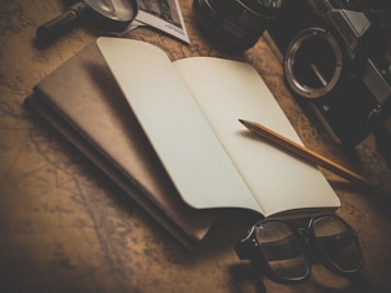 An open notebook lies on a vintage map with a pencil resting on it. Nearby, a pair of glasses, an old camera, and a magnifying glass can be seen, evoking a sense of exploration and creativity.