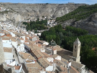 A picturesque village nestled in a rocky valley, showcasing whitewashed houses with terracotta roofs. A prominent church tower and dome stand out amidst the buildings. Lush greenery fills the valley against rugged, textured hillsides in the background.