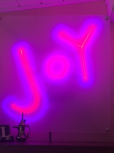 A vibrant neon sign displaying the word 'Joy' in bold, glowing letters. The sign is illuminated in bright pink and surrounded by a purple aura, creating a captivating visual effect. Below the sign, there is a decorative teapot-like object.