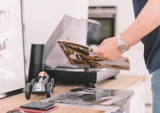 A person holding a vinyl record near a turntable. On the wooden surface, there is a toy car with the number 5 on it, a black cylindrical speaker, and more records. The setting appears to be a modern and tidy room, conveying a sense of music appreciation and leisure.