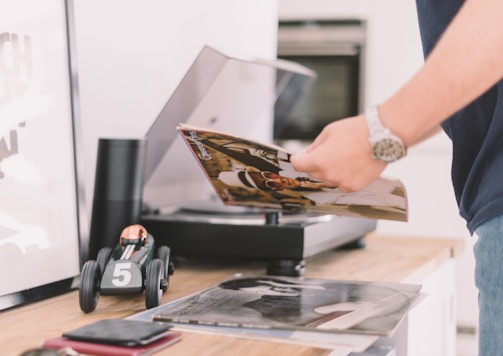 A person holding a vinyl record near a turntable. On the wooden surface, there is a toy car with the number 5 on it, a black cylindrical speaker, and more records. The setting appears to be a modern and tidy room, conveying a sense of music appreciation and leisure.
