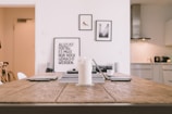 A minimalist and modern interior scene with a wooden table in the foreground. On the table, there are electronic devices, books, and a candle. In the background, framed pictures hang on a white wall, and there is a glimpse of a tidy kitchen with minimalistic design elements.