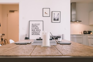 A minimalist and modern interior scene with a wooden table in the foreground. On the table, there are electronic devices, books, and a candle. In the background, framed pictures hang on a white wall, and there is a glimpse of a tidy kitchen with minimalistic design elements.
