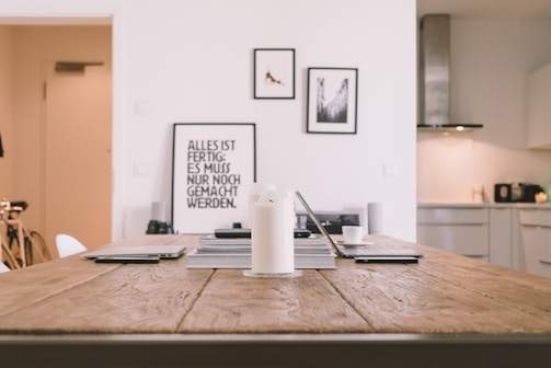 A minimalist and modern interior scene with a wooden table in the foreground. On the table, there are electronic devices, books, and a candle. In the background, framed pictures hang on a white wall, and there is a glimpse of a tidy kitchen with minimalistic design elements.