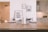 A minimalist and modern interior scene with a wooden table in the foreground. On the table, there are electronic devices, books, and a candle. In the background, framed pictures hang on a white wall, and there is a glimpse of a tidy kitchen with minimalistic design elements.