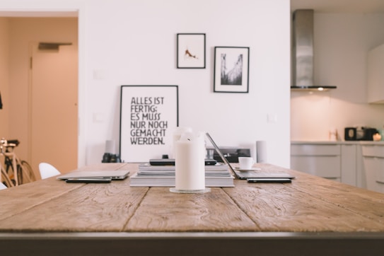 A minimalist and modern interior scene with a wooden table in the foreground. On the table, there are electronic devices, books, and a candle. In the background, framed pictures hang on a white wall, and there is a glimpse of a tidy kitchen with minimalistic design elements.