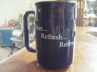 A black ceramic mug with the words 'Relax... Refresh... Renew.' written in white lettering. The mug is placed on a wooden table, and the background appears to be a cluttered interior space with indistinct objects such as shelves and furniture.