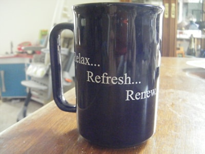 A black ceramic mug with the words 'Relax... Refresh... Renew.' written in white lettering. The mug is placed on a wooden table, and the background appears to be a cluttered interior space with indistinct objects such as shelves and furniture.