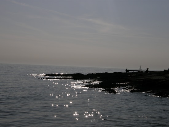 The image features a calm seascape with gentle waves and glittering reflections of sunlight on the water surface. A silhouetted shoreline with a few people is visible on the right. In the background, a small sailboat is seen against a grayish sky.