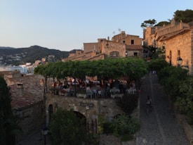 An outdoor restaurant with a leafy green canopy on a terrace overlooking a village. The stone buildings have rustic architecture. Hills and modern structures are visible in the background. The scene emits a serene and warm atmosphere as people appear to be enjoying a meal. The stone path leading up is lined with greenery.