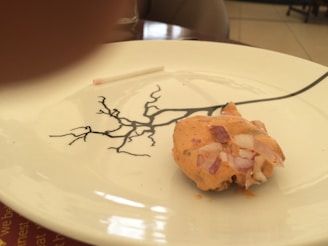 A piece of food garnished with chopped onions and a sauce is placed on a white plate with a decorative black branch design. The background is slightly blurred, indicating an indoor setting. A toothpick lies near the edge of the plate.